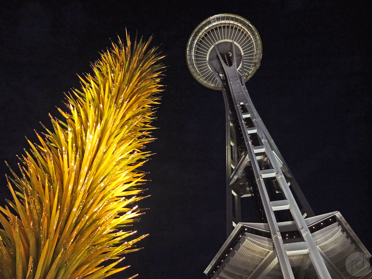 The Chihuly Garden and Glass exhibit at the Seattle Centre is illuminated at night with the Iconic Space Needle streching into the sky in the background.